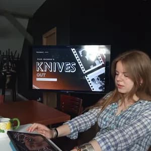 Młoda kobieta w koszuli w kratę patrzy na iPad przed telewizorem promującym film Riana Johnsona 'Knives Out'.