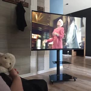 Widok z perspektywy pierwszej osoby z pluszową owieczką na kolanach i telewizorem wyświetlającym filmowy maraton w domowym salonie.