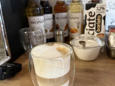 Szklanka z idealnie trójwarstwową kawą latte macchiato z gęstą pianką na górze. W tle na blacie stoją kolorowe syropy do kawy Monin oraz karton mleka.